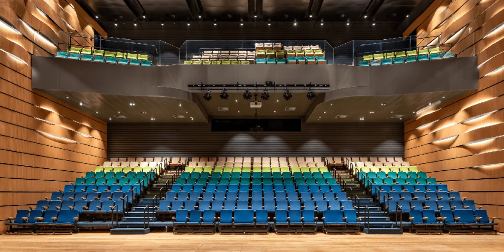 Visão geral de auditório empresarial contemporâneo: arquitetura corporativa com palco de madeira, poltronas em tons de azul e verde, mezanino e luminárias de palco, demonstrando expertise em design de espaços de eventos para corporações.