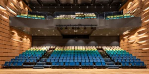Visão geral de auditório empresarial contemporâneo: arquitetura corporativa com palco de madeira, poltronas em tons de azul e verde, mezanino e luminárias de palco, demonstrando expertise em design de espaços de eventos para corporações.