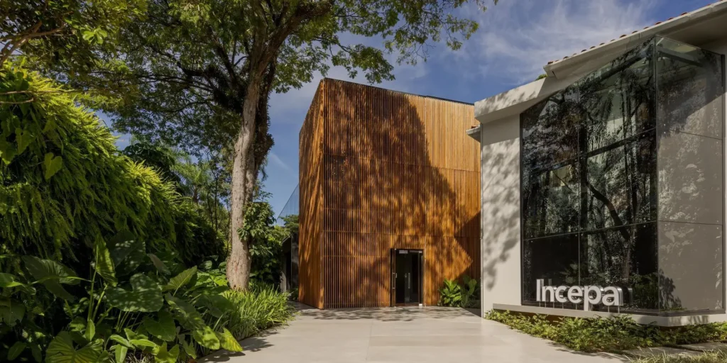 Fachada de escritório comercial moderno com arquitetura contemporânea, apresentando um cubo de madeira ripada e grande vidraça ao lado de um exuberante paisagismo tropical com samambaias e palmeiras, projetado por escritório de arquitetura.